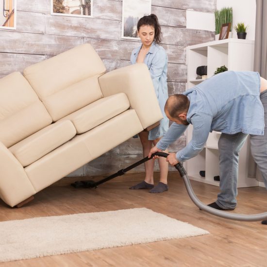 Wife picks up sofa Wife picks up sofa while her husband is cleaning the dust under it with vacuum cleaner.